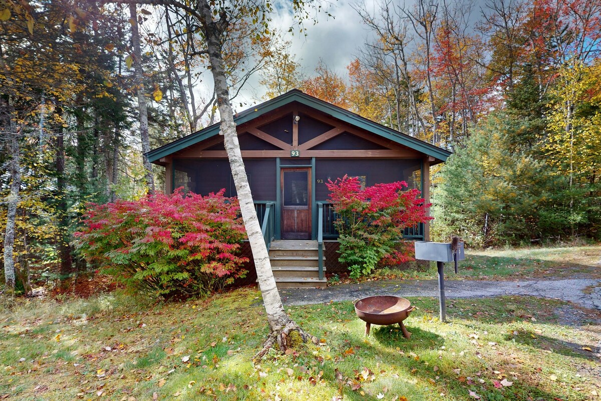 The cozy cabin exterior is framed by vibrant foliage in autumn hues. A set of steps leads to a front porch with a railing, while a firepit is visible to the side. The surrounding area features a lush lawn and trees, blending harmoniously with the natural landscape.