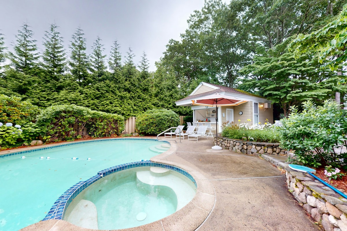 A private pool area is surrounded by lush greenery and landscaped gardens. An attached hot tub is visible next to the pool. Lounge chairs are arranged on the concrete patio beneath a shaded umbrella, providing a comfortable space for relaxation.