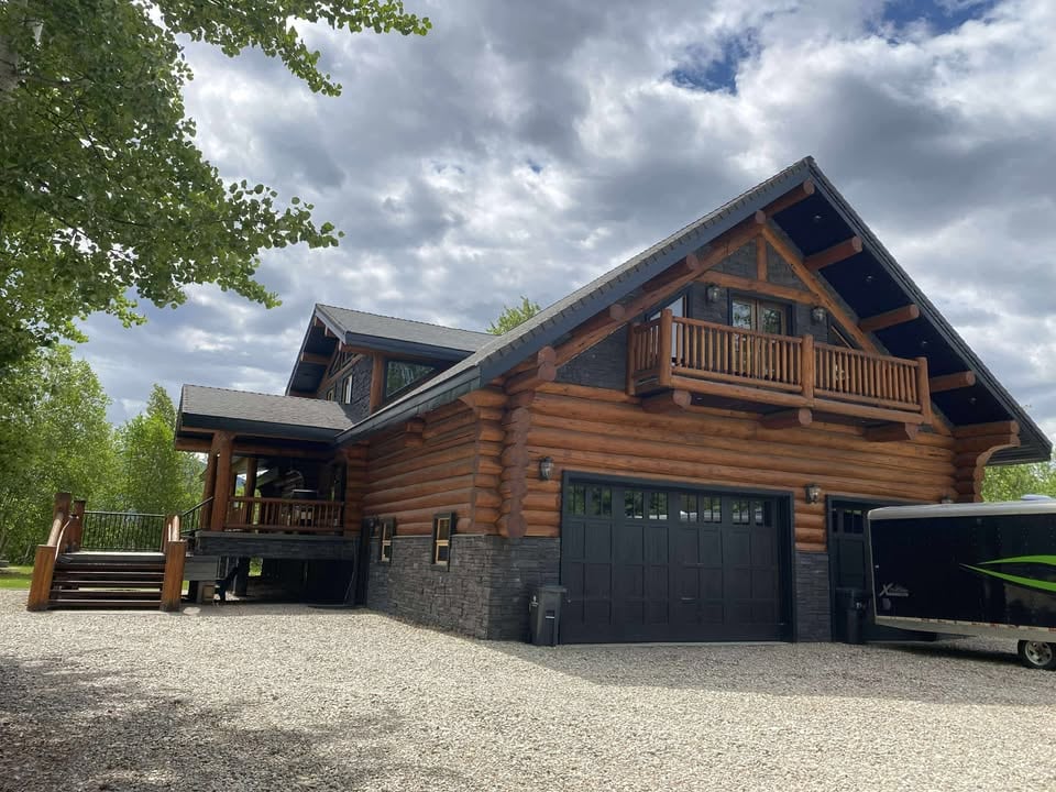 A large log cabin structure showcases a combination of natural wood and stone elements. The multi-level design features a prominent balcony and covered porch area. A gravel driveway leads up to the garage, framed by lush greenery under a partly cloudy sky.