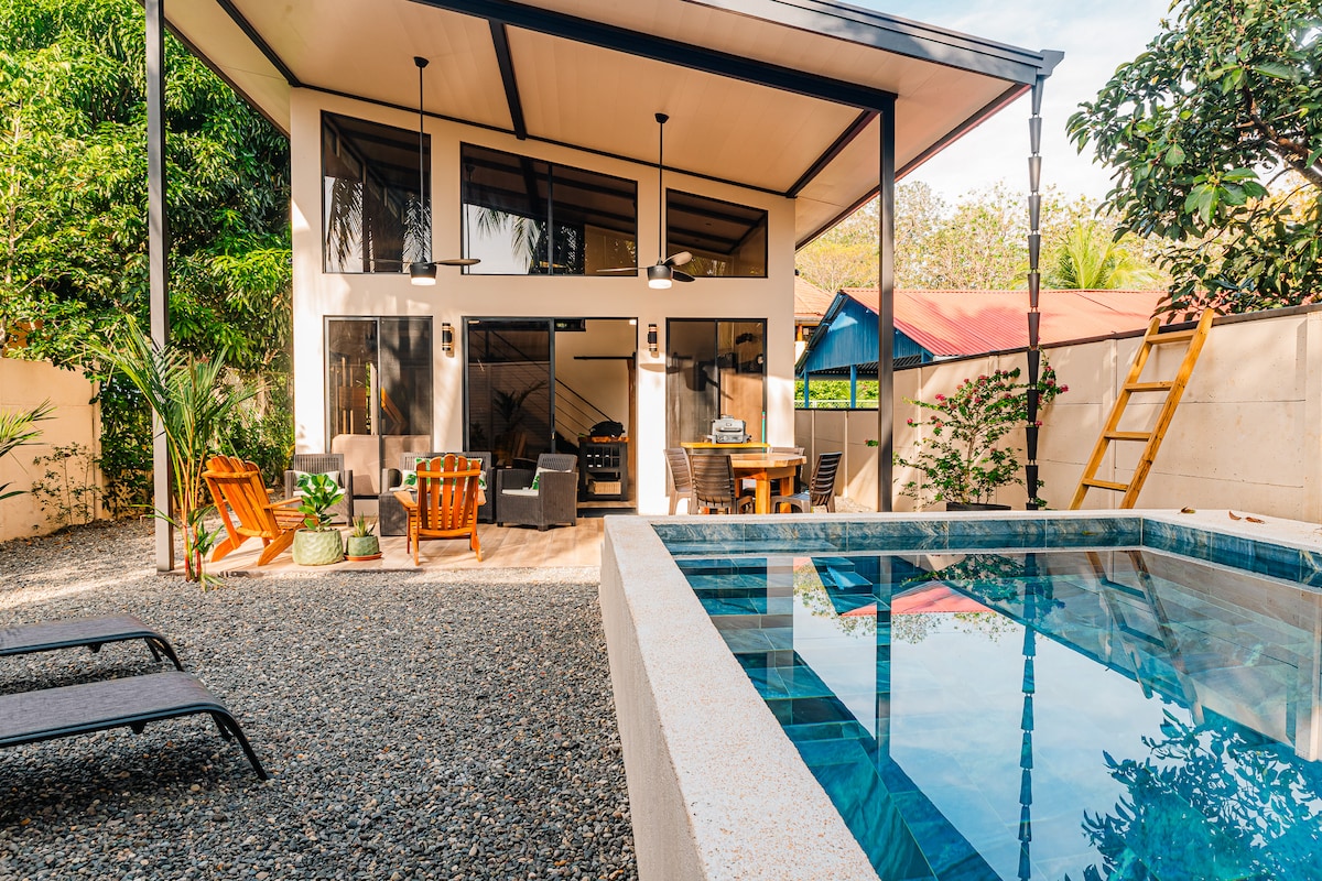 A modern villa is showcased from the poolside, featuring large glass doors that open to an inviting outdoor living area. Comfortable seating, a dining table, and tropical plants surround the private pool, offering a serene atmosphere under the bright blue sky.