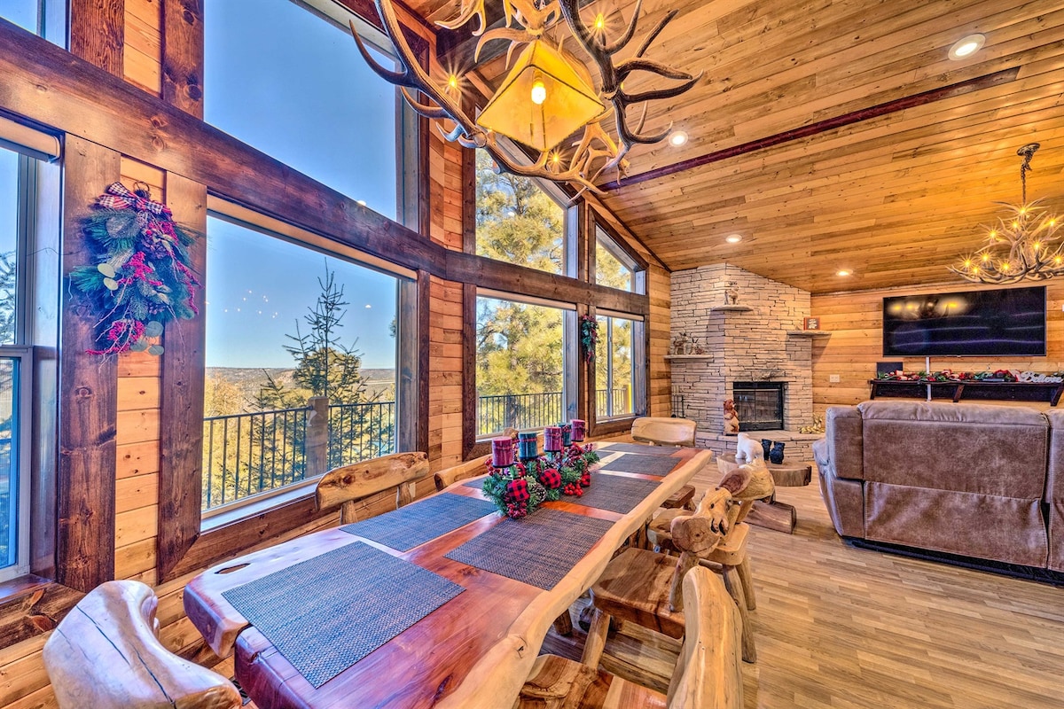 A spacious dining area features a long wooden table set with placemats, surrounded by rustic chairs. Large windows provide views of the surrounding landscape, while natural light illuminates the wood-paneled walls. A fireplace adds a cozy touch, complemented by decorative accents and overhead lighting.