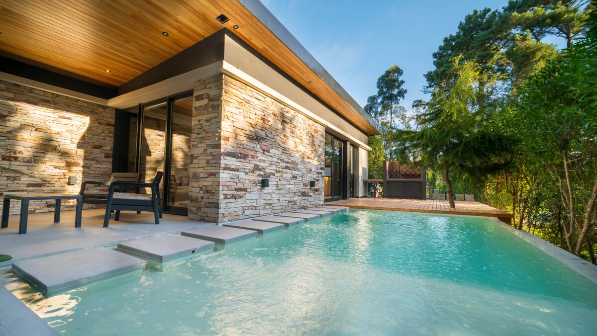 An outdoor area features a sparkling pool surrounded by smooth stone tiles and sunlit wooden decking. The building showcases a modern stone facade, with large glass doors opening to the pool area, allowing for a seamless transition between indoor and outdoor spaces.