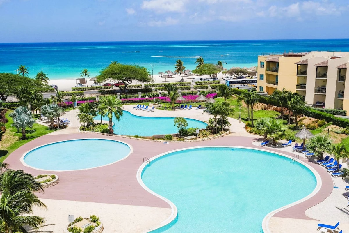 The image presents an expansive view of the resort's pools, featuring a series of circular shapes surrounded by well-maintained landscaping. Lush palm trees and vibrant flowering plants frame the area, with the stunning turquoise waters of Eagle Beach visible in the background.