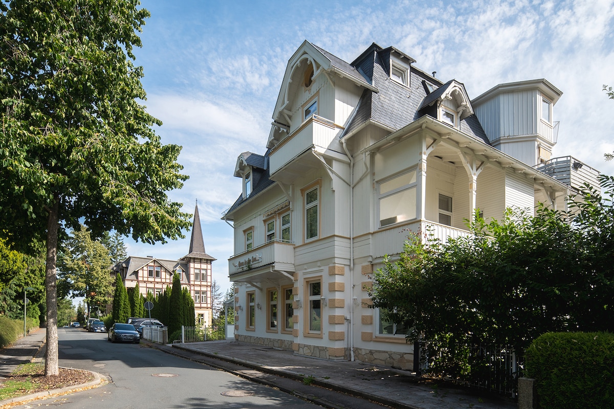 The exterior of a charming multi-story building is presented, featuring a mix of architectural elements. The façade is light-colored with decorative trim, surrounded by greenery. Nearby, a tree-lined street is visible, framed by additional buildings with varying styles in the background.
