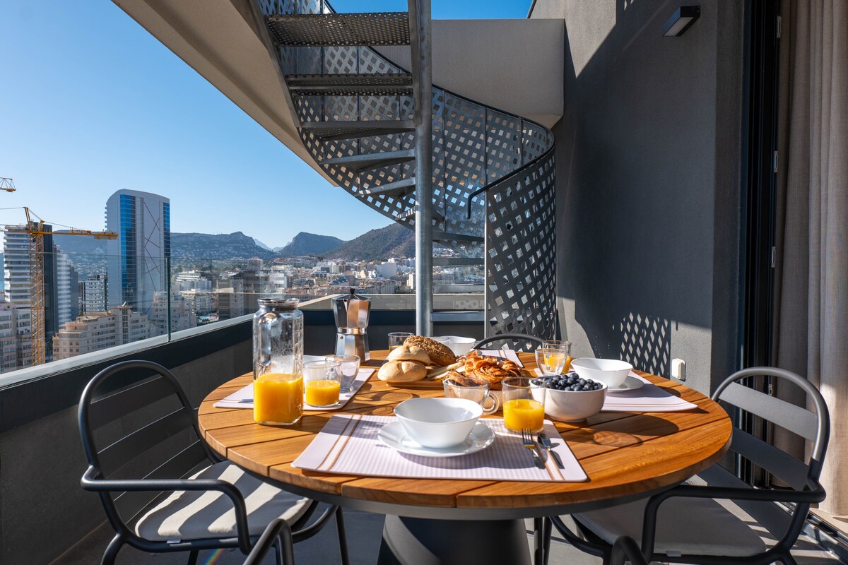 A round wooden dining table is set for breakfast on a spacious balcony. White dishes hold cereal and yogurt, accompanied by glasses of orange juice. The background features a stunning view of the city and mountains, with a spiral staircase leading to additional outdoor space.