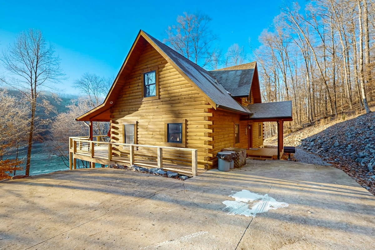 The exterior view of a spacious cabin is displayed, featuring a wooden construction with a high-pitched roof. A wraparound deck provides access to the outdoors, while the driveway offers ample parking space. Surrounding trees add to the natural setting near Norris Lake.