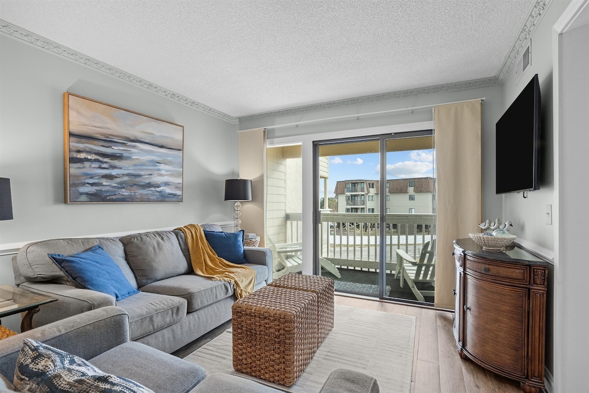 A comfortable living area is presented with a spacious sectional sofa adorned with blue and yellow pillows. Natural light enters through sliding glass doors leading to a private balcony, while a wooden coffee table enhances the space. A large artwork piece complements the soft color scheme.