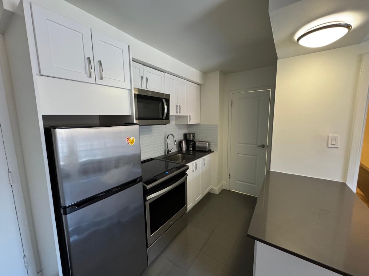 A modern kitchen is showcased with sleek cabinetry and a neutral color palette. Stainless steel appliances, including a refrigerator, microwave, and stove, are neatly arranged. A countertop provides ample prep space, contributing to a well-equipped cooking environment.