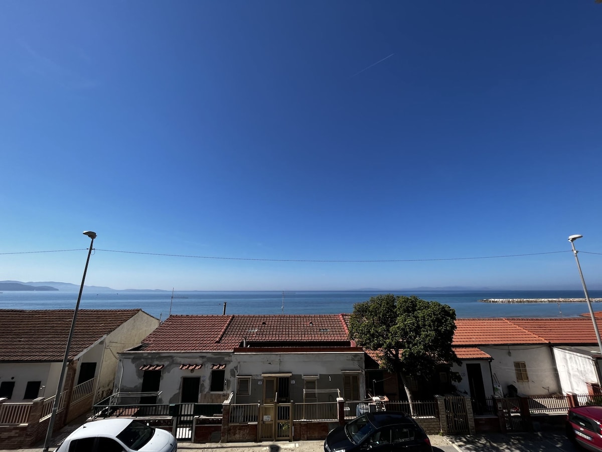 A clear blue sky stretches over a calm sea, visible across the horizon. The image captures a row of low structures with red-tiled roofs lining the foreground, creating a coastal seaside atmosphere. The peaceful setting is accentuated by the absence of clouds.