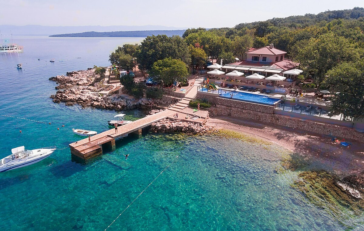 An aerial view captures a coastal setting featuring crystal-clear waters and a sandy beach. Sun loungers line the pool area, while a wooden pier extends into the water. Nearby, trees provide shade, and various boats are visible on the horizon.