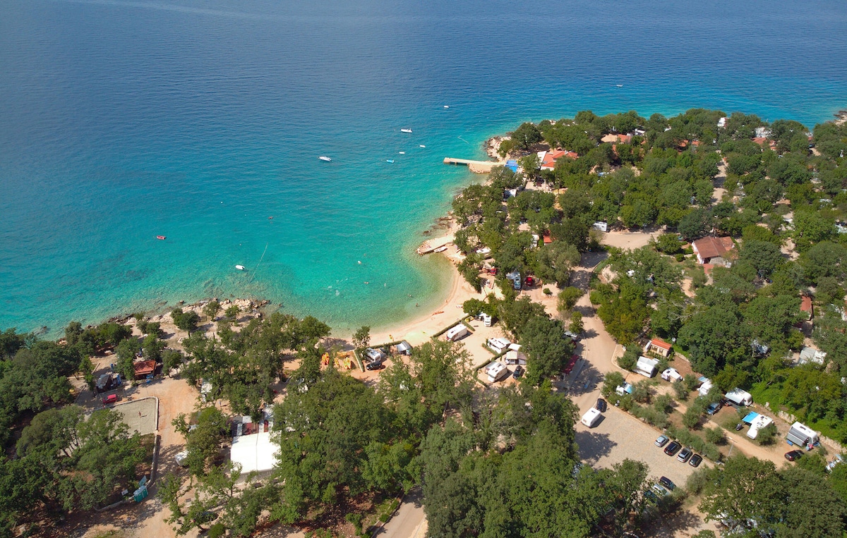 An aerial view captures a serene beach with clear turquoise waters. The surrounding area is dotted with lush trees and several accommodations. Small boats can be seen in the water, and sandy shores invite relaxation. A parking area is visible, along with grassy spaces for outdoor activities.