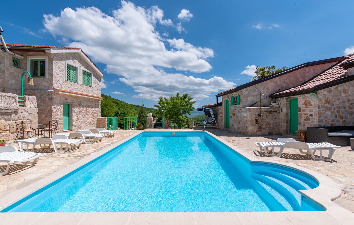 An outdoor private pool is featured, surrounded by sun loungers and stone-paved areas. Lush greenery is visible in the background, and stone structures with green shutters add charm to the setting under a bright blue sky with scattered clouds.
