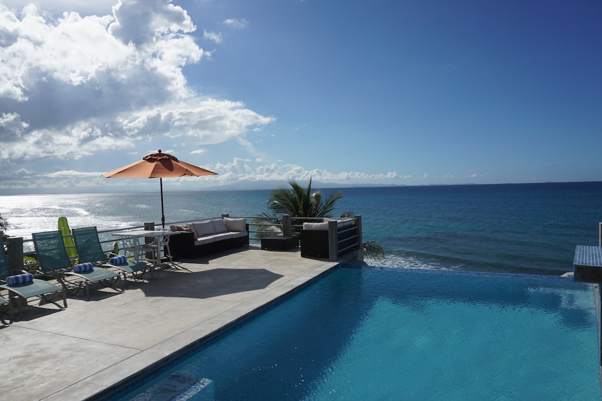 An infinity pool extends toward the ocean, featuring a natural stone waterfall. Lounge chairs with striped cushions are arranged around the pool area. A large umbrella provides shade over a sitting area with sofas facing the water, while palm trees are visible in the background.