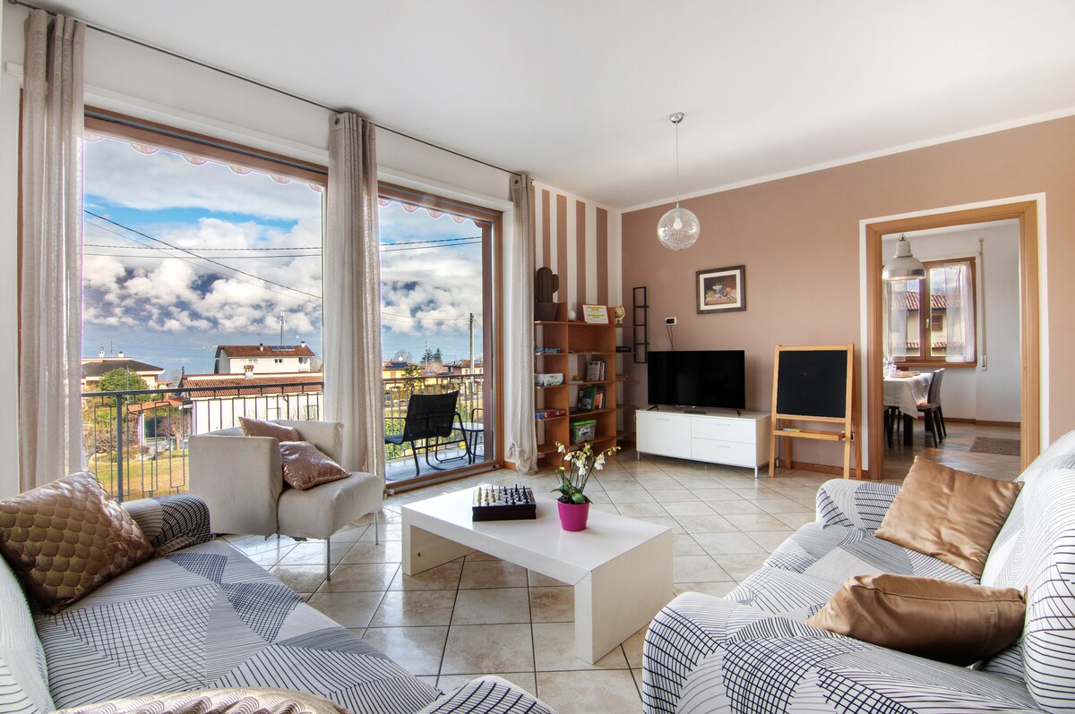 The living room features large windows offering views of the surrounding landscape. Comfortable seating is arranged around a coffee table, while a TV and bookshelf are positioned nearby. A dining area is accessible through an archway, enhancing the open layout of the space.