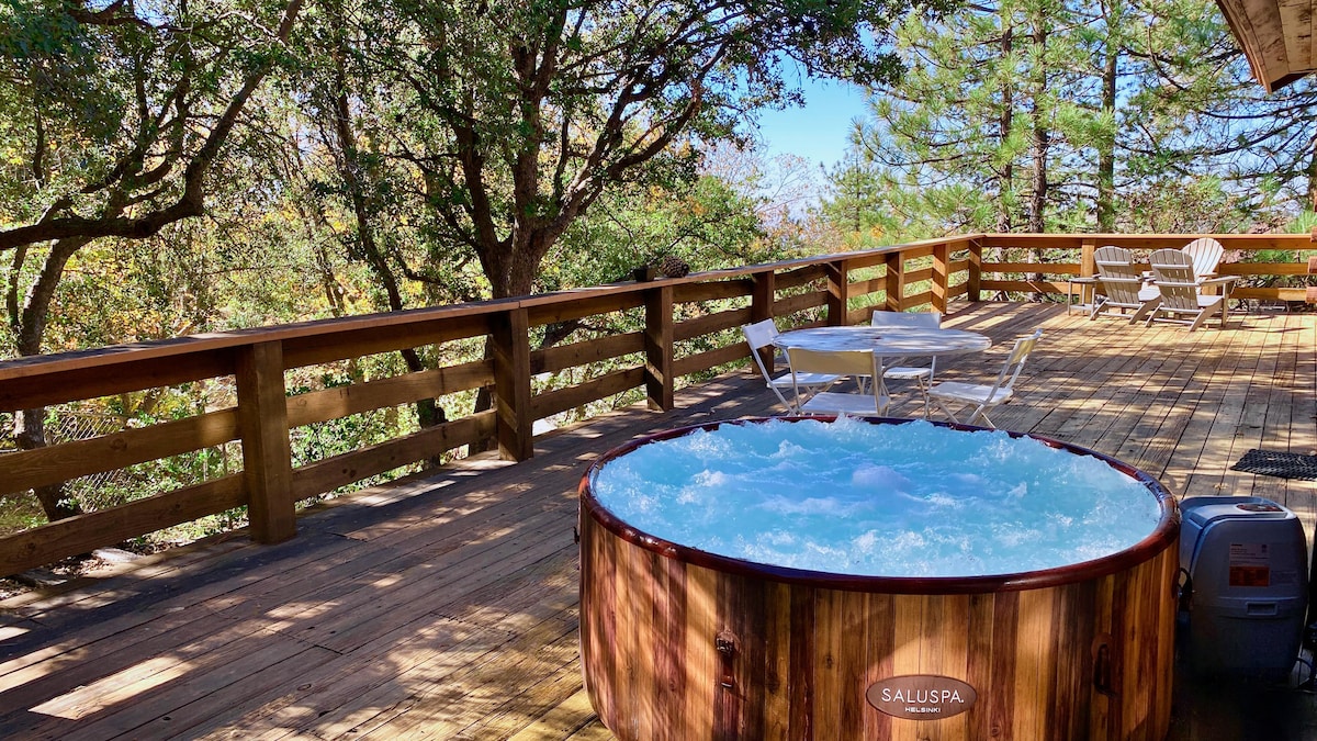 An expansive wooden deck is featured, surrounded by greenery. A round hot tub is positioned close to seating arrangements, including a table and chairs, inviting relaxation amidst nature. Sunlight filters through the trees, creating a serene outdoor environment.