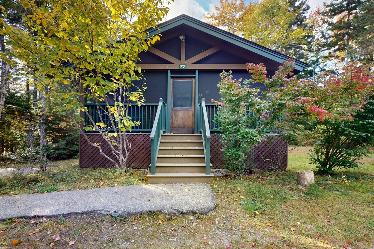 A view of the Tamarack Cabin features a welcoming entrance with a sturdy set of stairs leading to a covered porch. Surrounding trees and colorful shrubs add natural beauty to the cabin's exterior, creating a serene setting in the woods.
