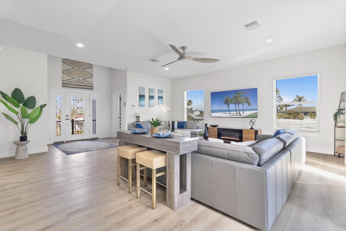 A spacious living area features large windows that allow natural light to fill the room. A comfortable sectional sofa is arranged near a modern entertainment center, while a wooden dining table with two stools adds functionality. Decorative plants enhance the welcoming environment.