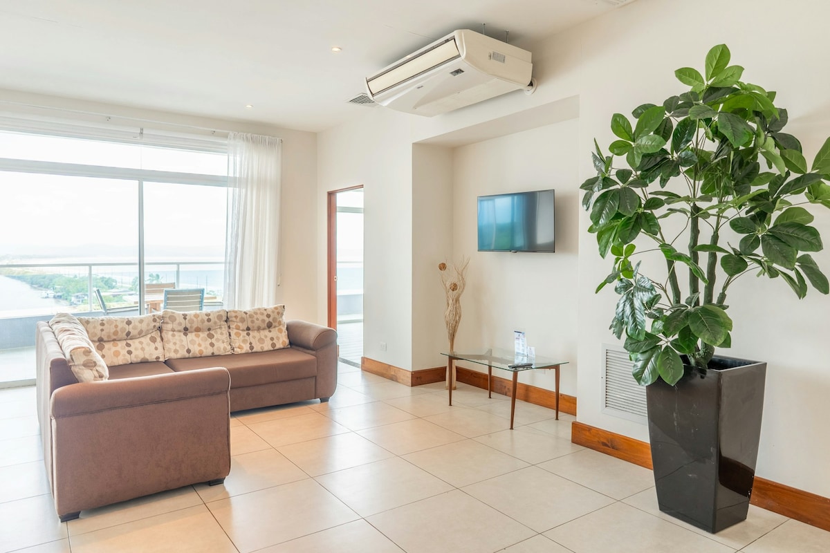 A spacious living area features a comfortable brown sofa adorned with patterned cushions. A flat-screen TV is mounted on the wall, with a sleek glass coffee table placed in front. A large window offers views of the ocean and surrounding landscape, with a potted plant adding a touch of greenery.