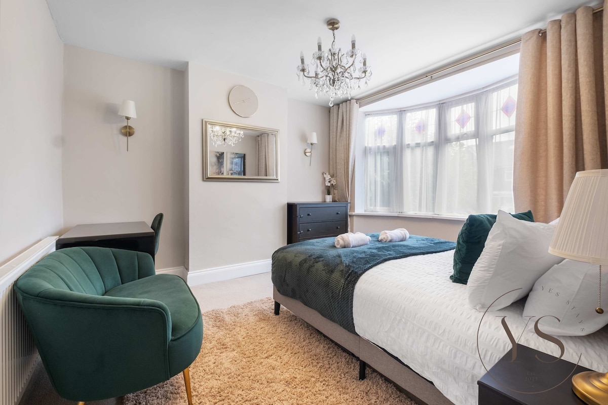 A comfortable bedroom features a spacious bed with plush bedding and decorative pillows. A stylish armchair and a small desk are present, alongside a sleek dresser. Natural light filters through large windows adorned with elegant curtains, creating a bright and welcoming environment.