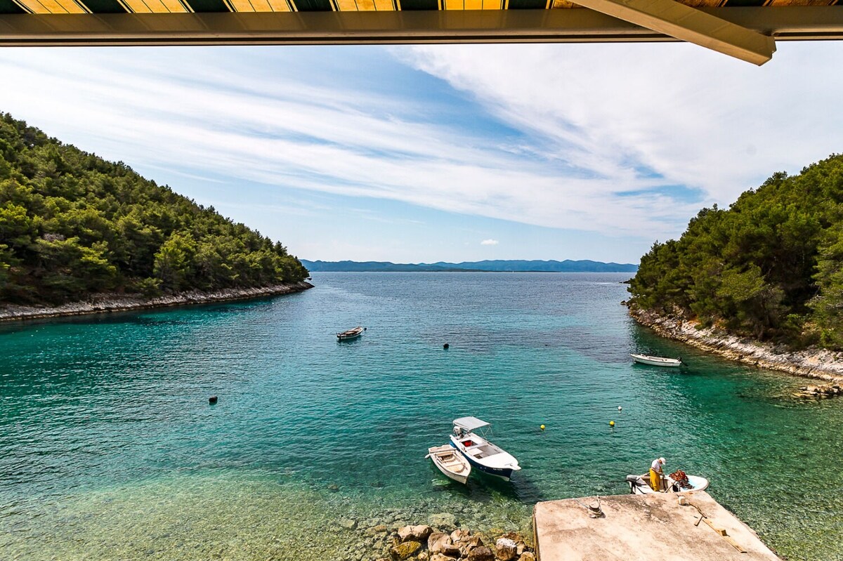 A scenic view of a calm turquoise bay is captured, with small boats gently anchored in the clear water. Lush green trees line the shore, and the horizon features distant islands under a partly cloudy sky.