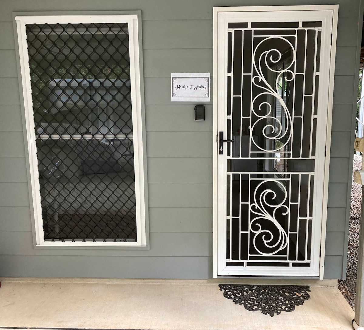 A private entrance is showcased through an intricately designed security door with a decorative pattern. Adjacent is a large window secured with a grid, providing natural light while ensuring safety. The welcome sign is displayed to enhance the inviting entrance.