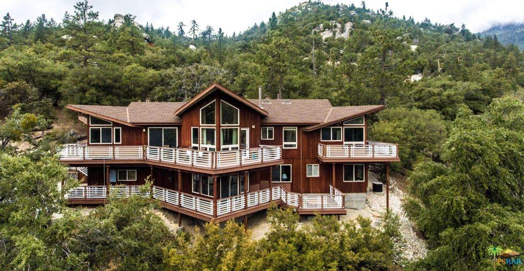 A large multi-level cabin with a wooden exterior is surrounded by greenery, showcasing multiple balconies and a prominent central window. The architecture integrates with the natural landscape, highlighting the mountain backdrop and a serene forest setting.
