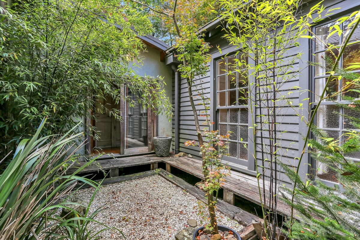 A serene garden area is presented with wooden decking and a gravel path. Small plants and bamboo foliage surround a central potted tree, adding natural elements to the space. A doorway leads into a cottage interior, blending outdoor and indoor environments harmoniously.