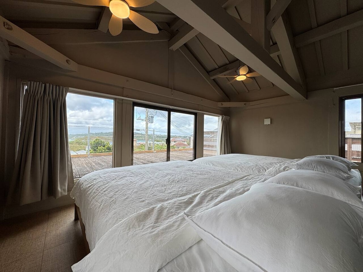 A spacious sleeping area features two double beds with crisp white bedding, softly illuminated by natural light from large windows. A scenic view of the surrounding landscape can be seen through the glass, while ceiling fans add a touch of comfort to the space.