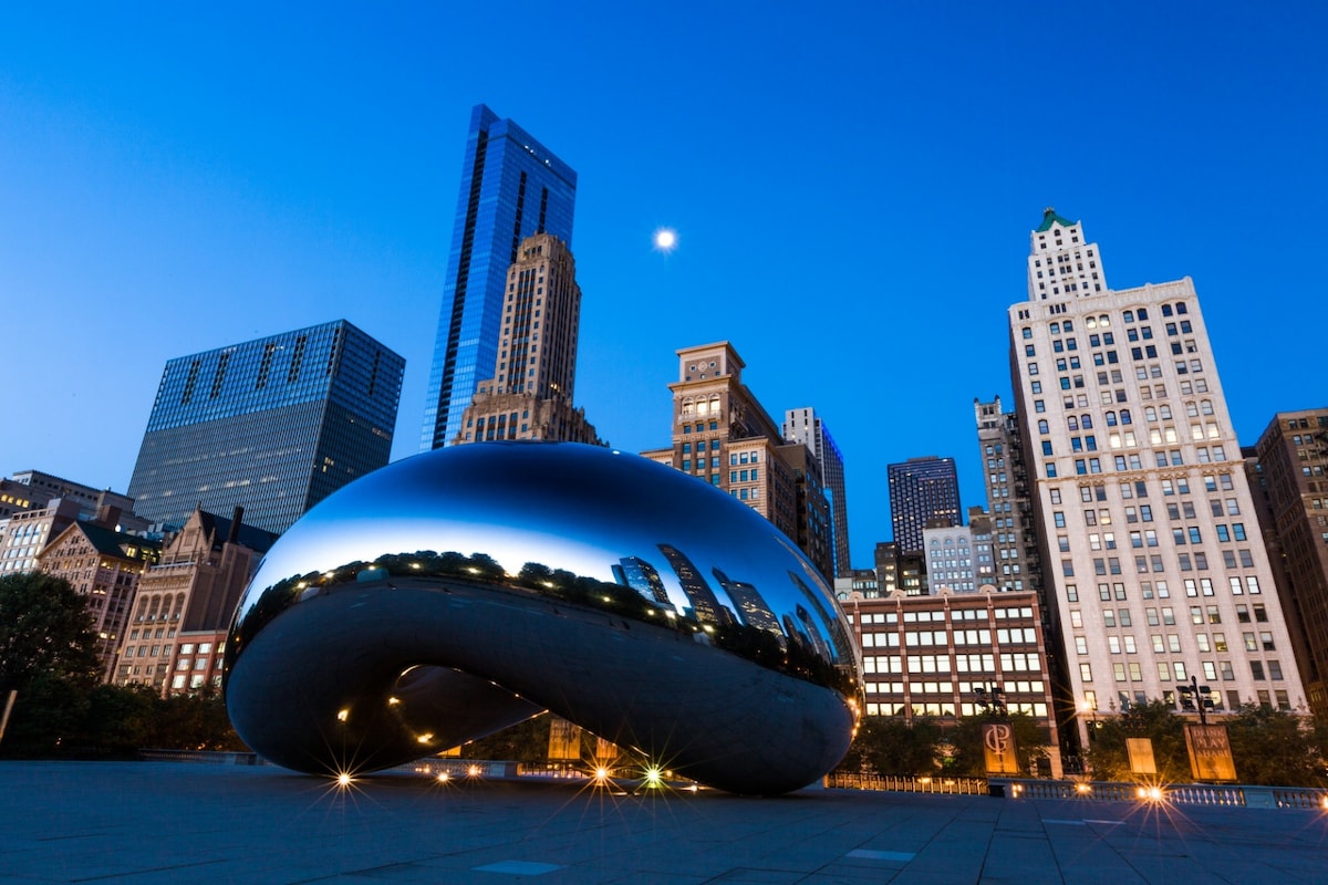 A large, reflective sculpture, known as 'The Bean,' is prominently displayed at Millennium Park, surrounded by an array of tall buildings. The evening sky casts a soft blue hue, with lights illuminating the sculpture and creating striking reflections of the city skyline.