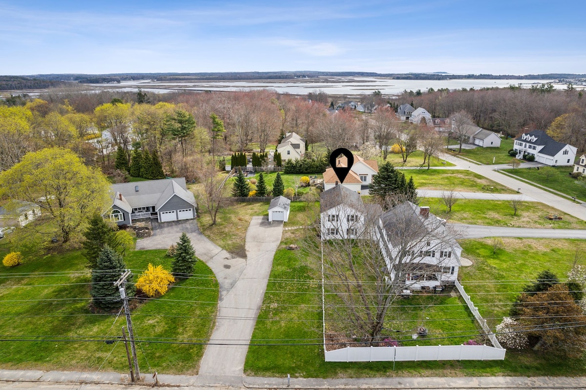 An aerial view captures the surrounding neighborhood and landscape, showcasing multiple homes set amidst open green spaces and trees. The river and marshland are visible in the background, providing a scenic backdrop to the residential area.