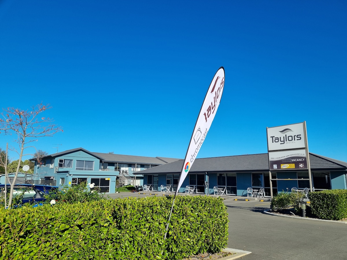 A modern building is displayed under a clear blue sky, featuring a sign indicating 'Taylors' with a vacancy notice. Outdoor seating areas with tables and chairs are visible in front, surrounded by well-maintained greenery.