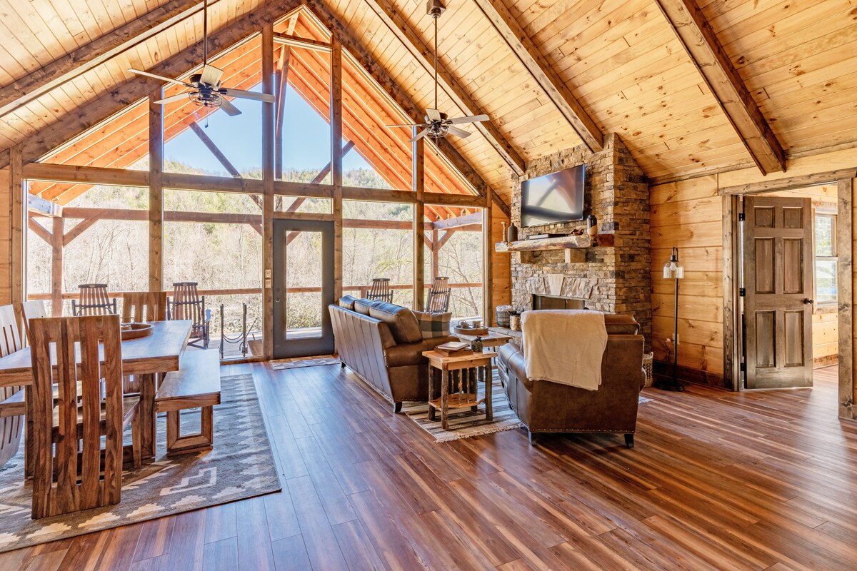 An open living space features wooden beams and a vaulted ceiling, with large windows allowing natural light and views of the outdoors. A comfortable seating area is positioned next to a stone fireplace, while a dining table accommodates multiple guests. Hardwood flooring enhances the inviting design.