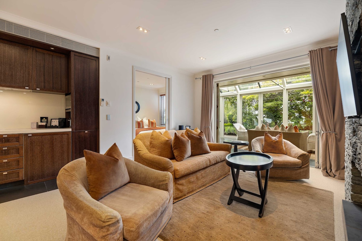 An inviting living area features plush brown sofas arranged around a circular table, with soft accent pillows providing comfort. Natural light floods in from the glass conservatory, illuminating the space and showcasing views of the surrounding greenery.