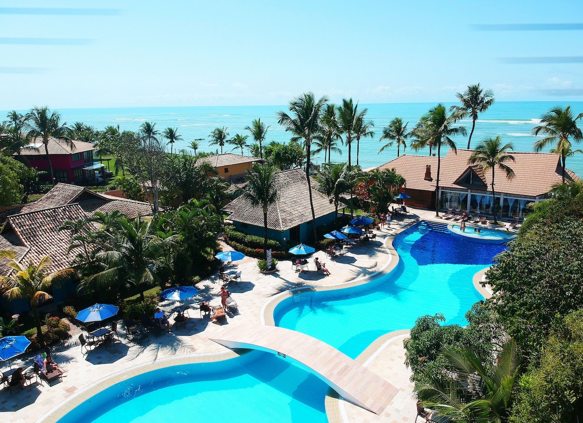An expansive view of a resort-style pool area is outlined by lush palm trees and vibrant umbrellas. The pool features winding shapes, leading toward the ocean in the background, where the water meets the sky. Soft lounge chairs are positioned around the pool for relaxation.