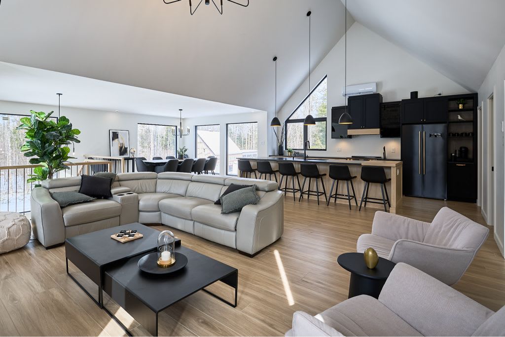 An open-concept living area features a modern, light-filled design with high ceilings and large windows. A sectional sofa and armchairs provide comfortable seating around a coffee table. The adjacent kitchen, with a dark color scheme, includes a dining area with barstools, creating a welcoming space for gathering.