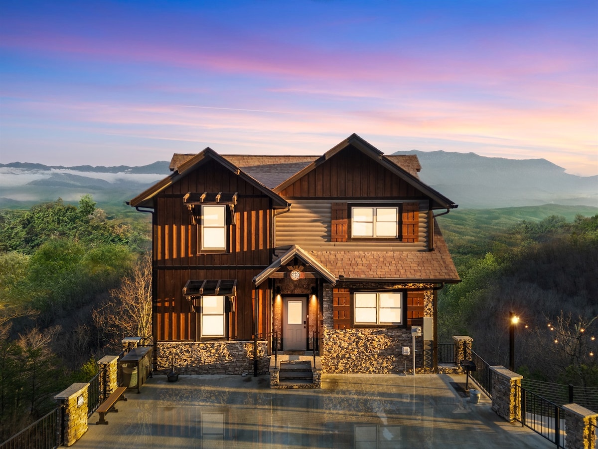 The exterior of a modern cabin showcases a blend of wood and stone materials, standing proudly against a backdrop of rolling mountains and an early morning sky. A welcoming entryway is featured, with a paved pathway leading to the front door.