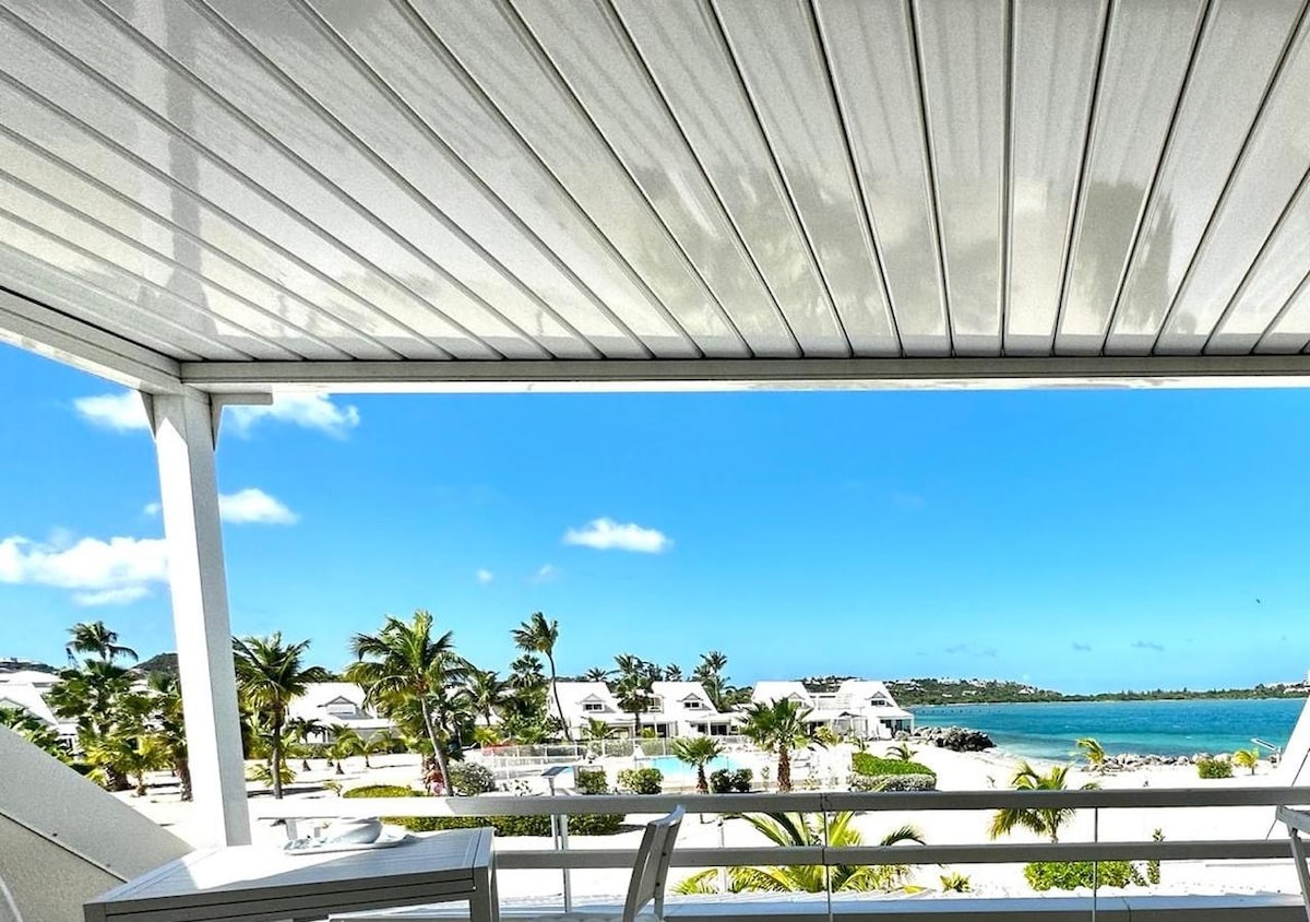 A shaded terrace offers a serene view of palm trees and the clear waters of the Caribbean Sea. The horizon displays a bright blue sky dotted with a few clouds, enhancing the peaceful beach setting. A table is set, inviting relaxation and enjoyment of the surroundings.