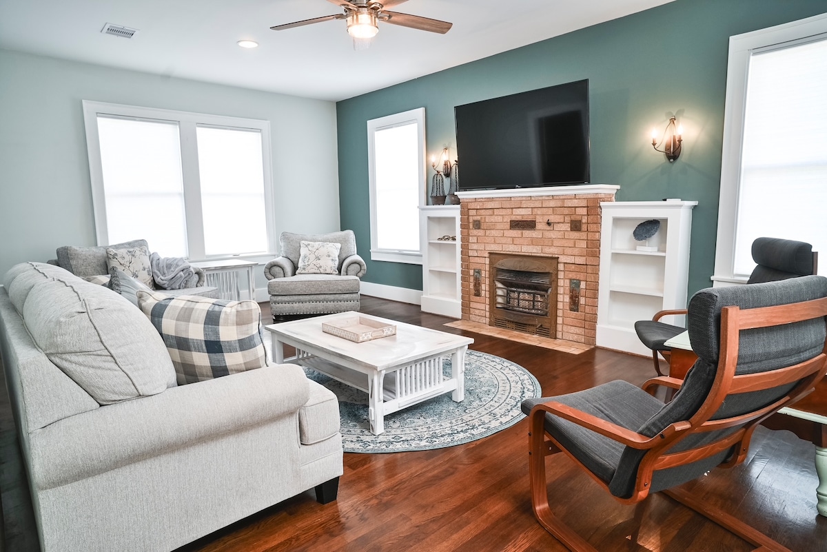 A comfortable living area is arranged with a light-colored sofa and patterned armchair, complemented by a mid-century style lounge chair. A wooden coffee table sits atop a round area rug, with a television mounted above a brick fireplace. Natural light enters through multiple windows.