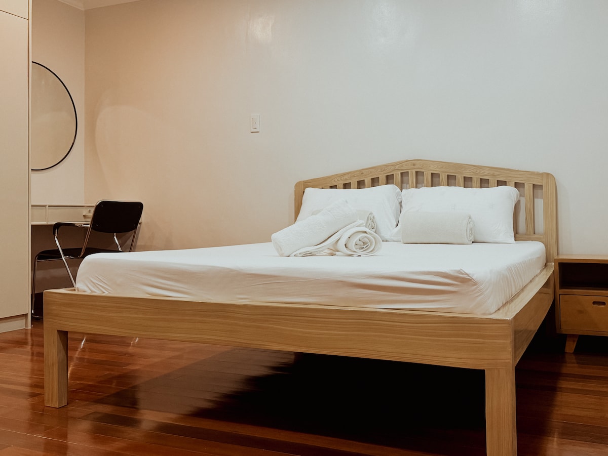 A spacious bed with clean white linens is positioned in a well-lit room. Soft towels are neatly arranged at the foot of the bed. A simple chair and desk are visible nearby, and soft wooden flooring enhances the calming ambiance.