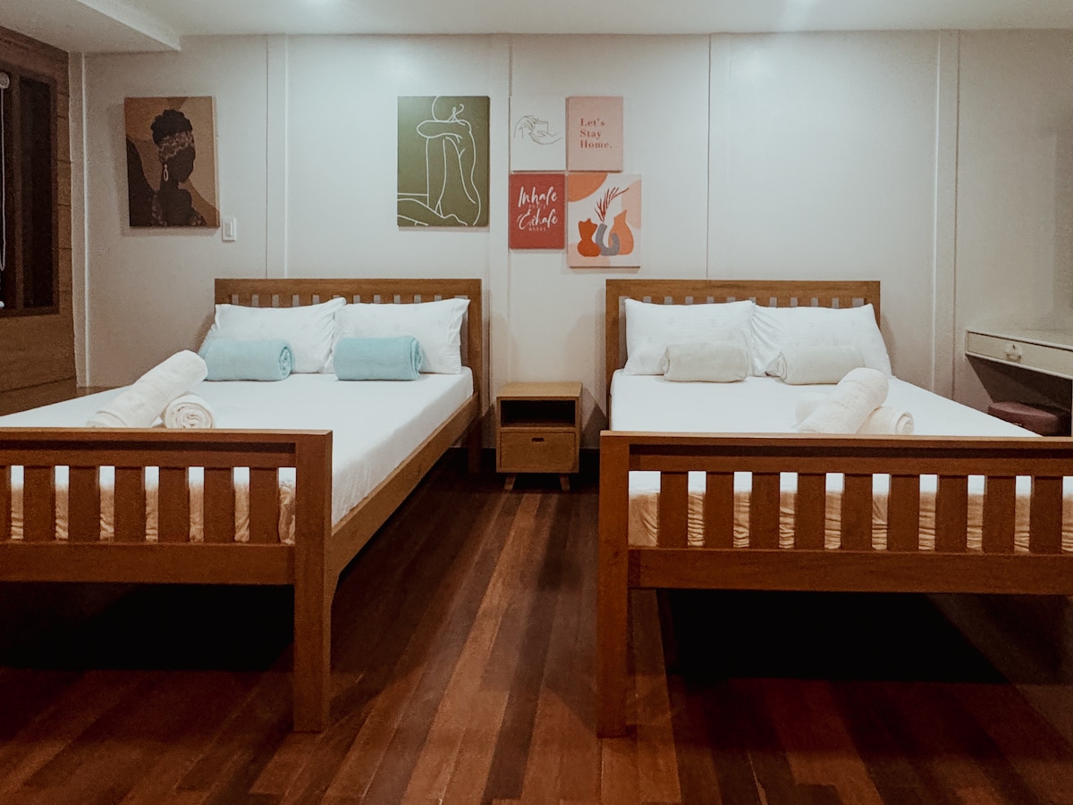 The room contains two double beds with wooden frames, each topped with white linens and light blue accent pillows. A nightstand is placed between the beds. Art pieces adorn the walls, and wooden flooring enhances the cozy atmosphere.