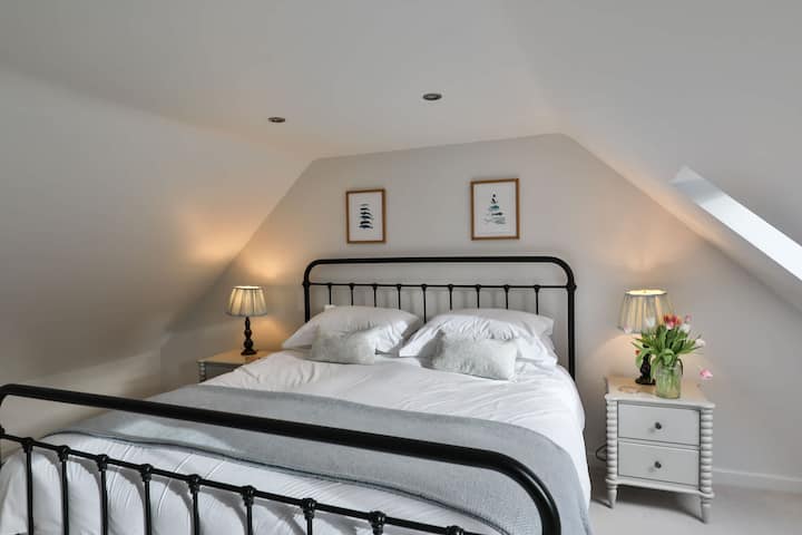 Desconecta con estilo en el dormitorio principal, bellamente decorado, con una lujosa cama tamaño king, sábanas blancas y tonos neutros relajantes. Los techos inclinados y la iluminación suave crean un refugio tranquilo para un descanso profundo