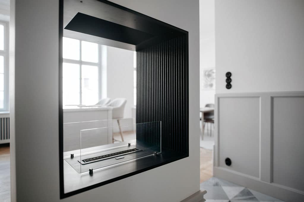 A modern electric fireplace is framed by sleek black panels, showcasing vertical slats that enhance its design. Soft light from surrounding windows illuminates the space, providing a warm focal point in the apartment's open layout.