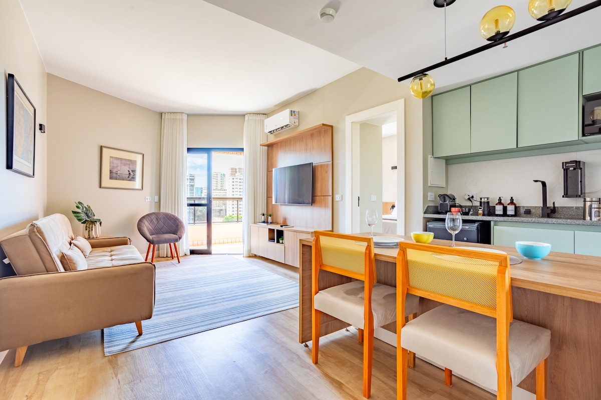 A spacious living area is showcased, featuring a cozy seating arrangement with a sofa and armchair. A dining table with two chairs sits in the foreground. Light-colored walls are complemented by large windows, allowing natural light to illuminate the space. Modern kitchen cabinetry is visible in the background.