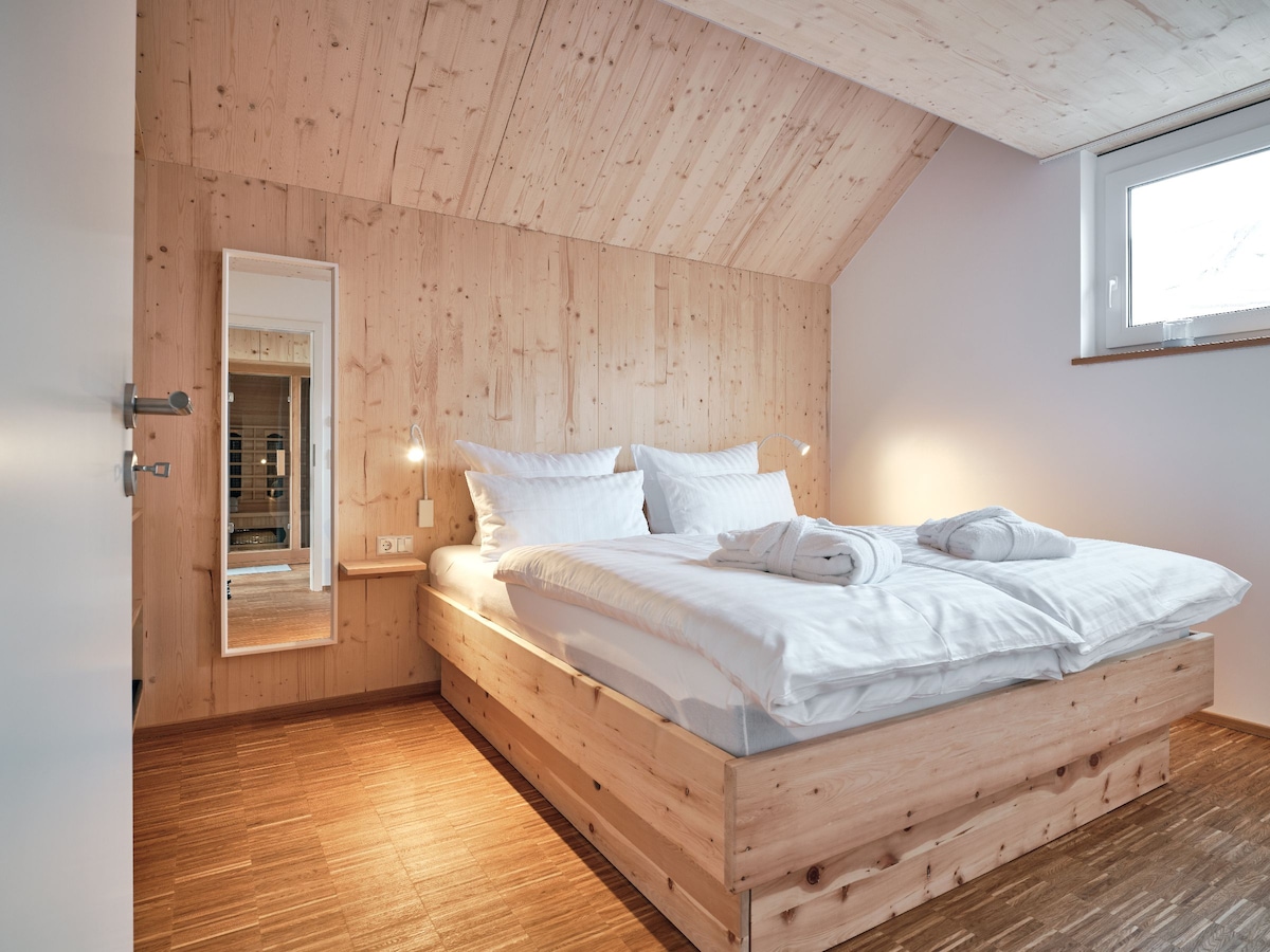 A bright bedroom is presented, featuring a spacious bed with crisp white linens and multiple plush pillows. The room is accented by warm wooden paneling and a mirror mounted on the wall. Natural light streams through a nearby window, creating a serene ambiance.