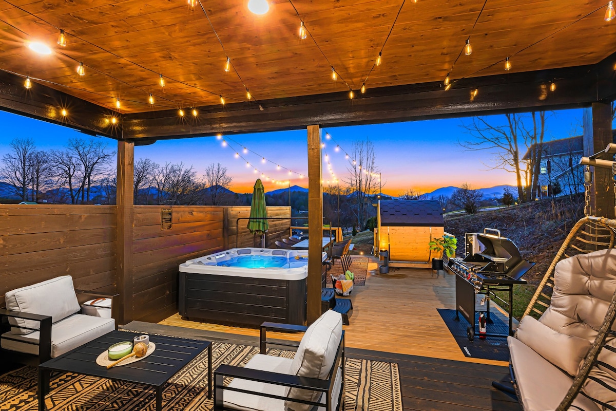 An outdoor entertainment area is featured, showcasing a hot tub beneath string lights. Comfortable seating arrangements are present, complemented by a barbecue grill and an additional shaded area. The backdrop displays a scenic view of distant mountains during sunset.