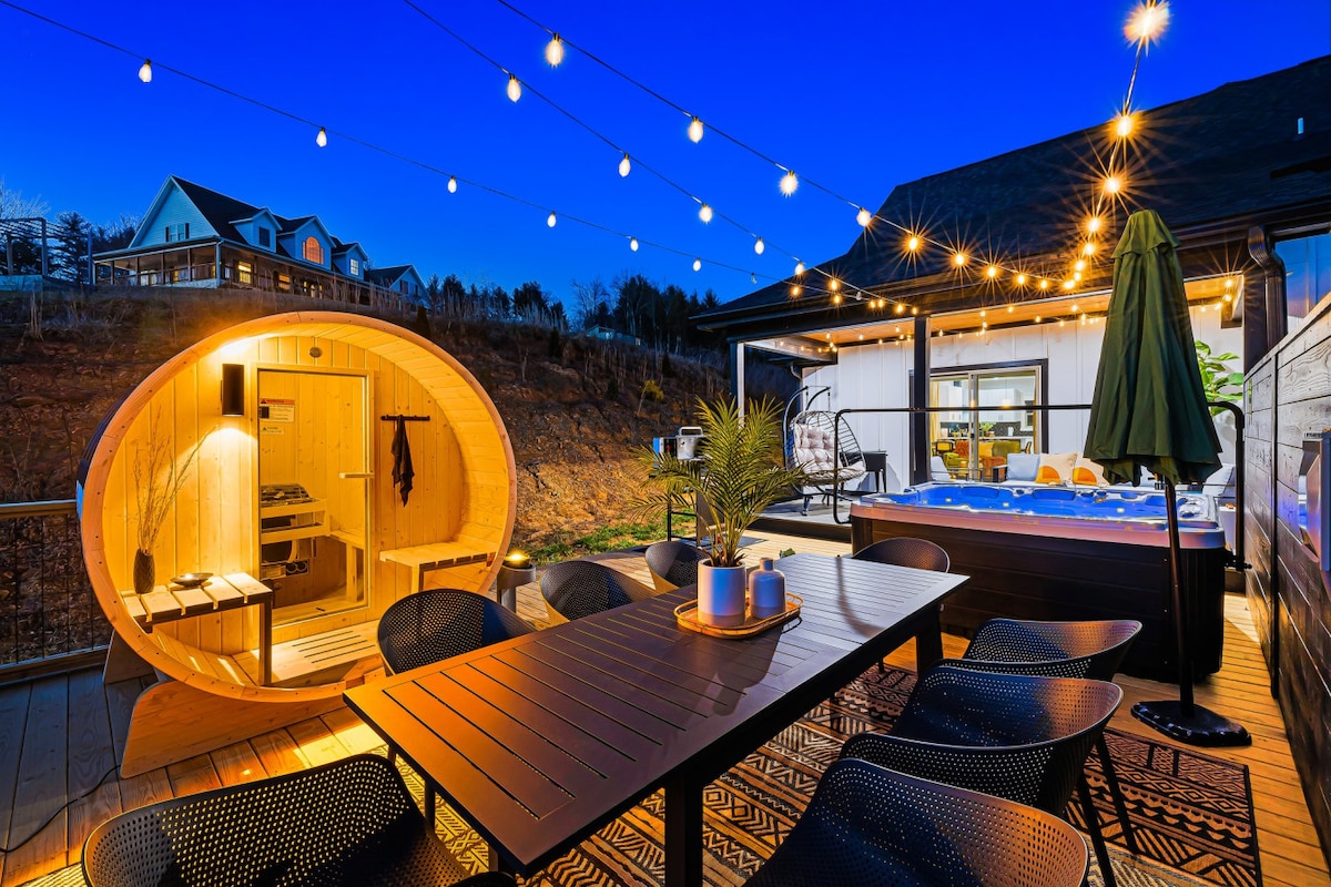 An outdoor dining area is visible, featuring a large table surrounded by black chairs. A private barrel sauna is positioned nearby, illuminated warmly. String lights hang above, adding a soft glow to the scene, while a hot tub is seen in the background against a twilight sky.