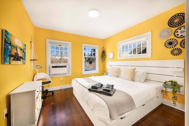 Un'accogliente camera da letto con pareti di colore giallo brillante, un telaio del letto in legno bianco con biancheria da letto neutra e opere d'arte murali con intricati disegni intrecciati. La stanza ha luce naturale da più finestre e una piccola zona lavoro con sedia.