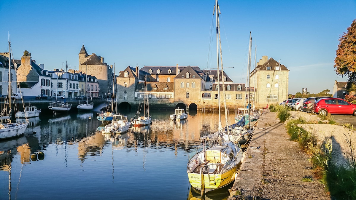 The image captures a serene harbor scene with several boats moored in calm waters. Historical stone buildings line the shore, reflecting the soft blue sky. Gentle ripples create reflections on the water's surface, contributing to the tranquil atmosphere of the setting.