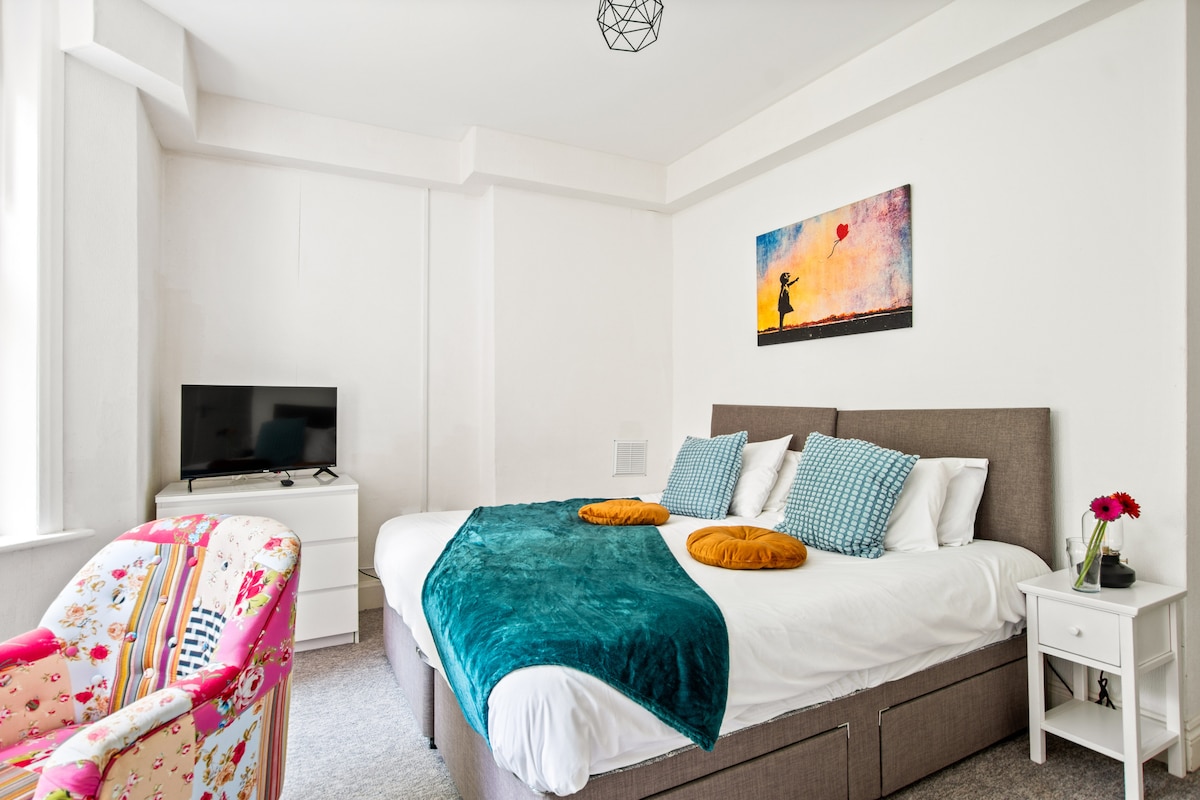 A spacious bedroom features a large bed adorned with colorful pillows and a soft throw. A vibrant piece of wall art hangs above the bed, and a small television is placed on a dresser. A cozy chair with a floral pattern complements the decor.