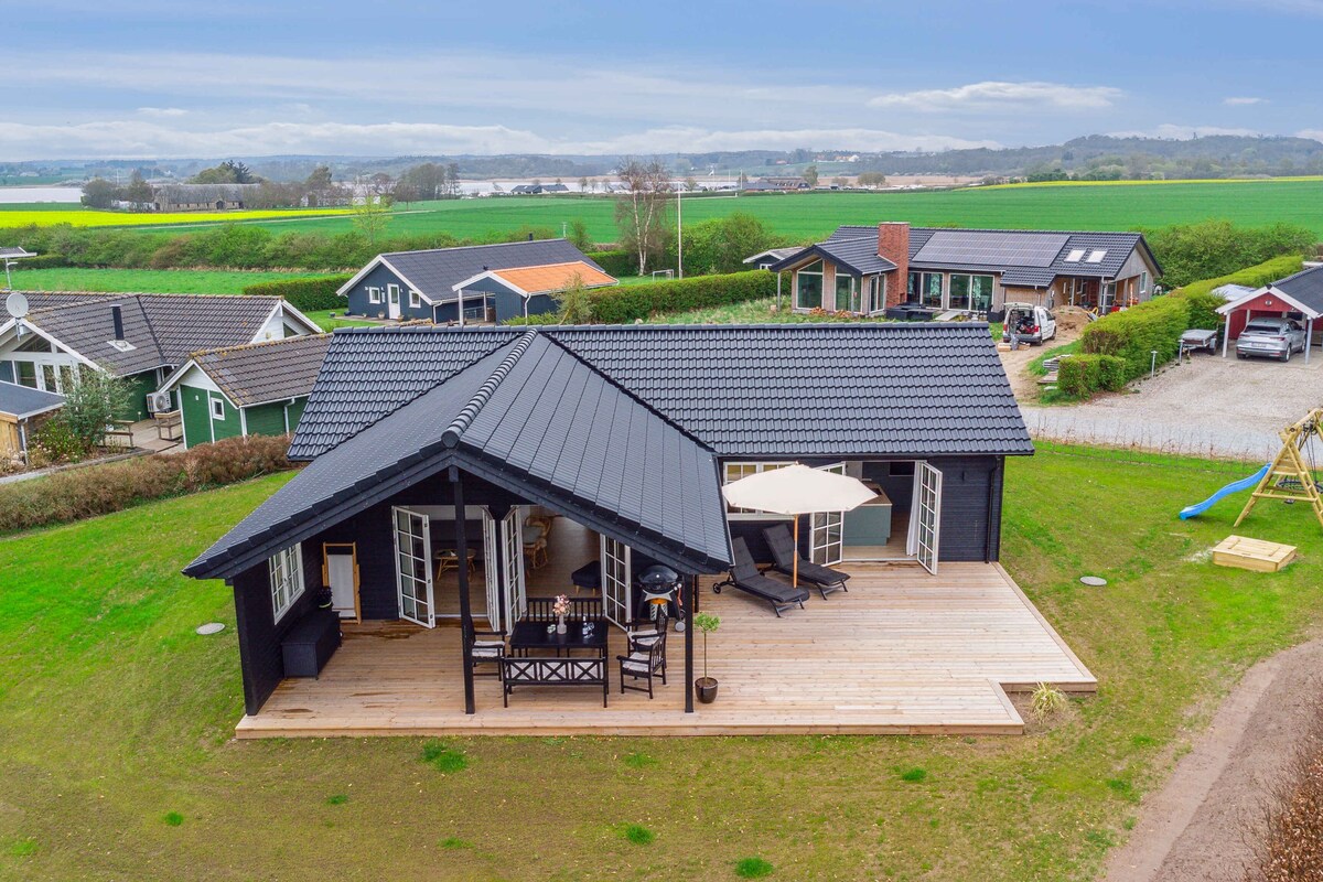 An aerial view of the modern holiday home showcases a spacious wooden terrace, furnished with a shaded seating area and lounge chairs. The surrounding green fields and neighboring houses create a peaceful backdrop, highlighting the home's inviting outdoor space.